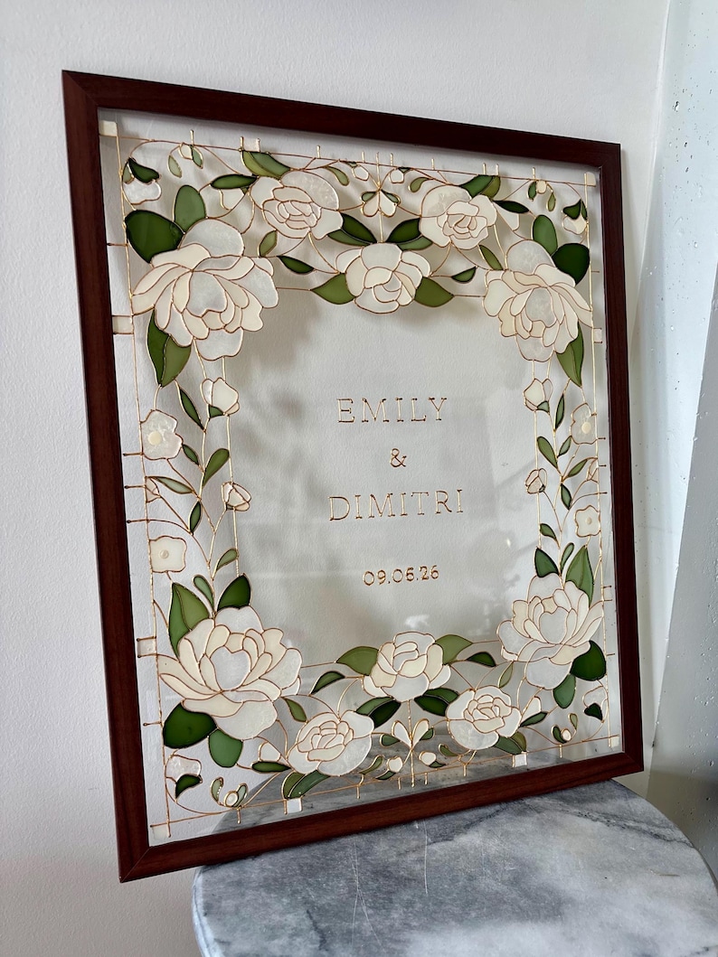 May include: A framed artwork featuring white floral designs and the names "EMILY & DIMITRI" with the date "09.05.26". The frame is a dark brown color, and the artwork has a transparent background.