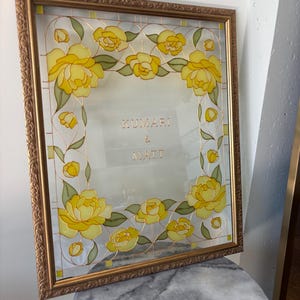May include: A framed glass art piece with a gold-colored frame. The glass features a floral design with yellow roses and green leaves. The names "KUMARI & MATT" are displayed in gold lettering within the frame.