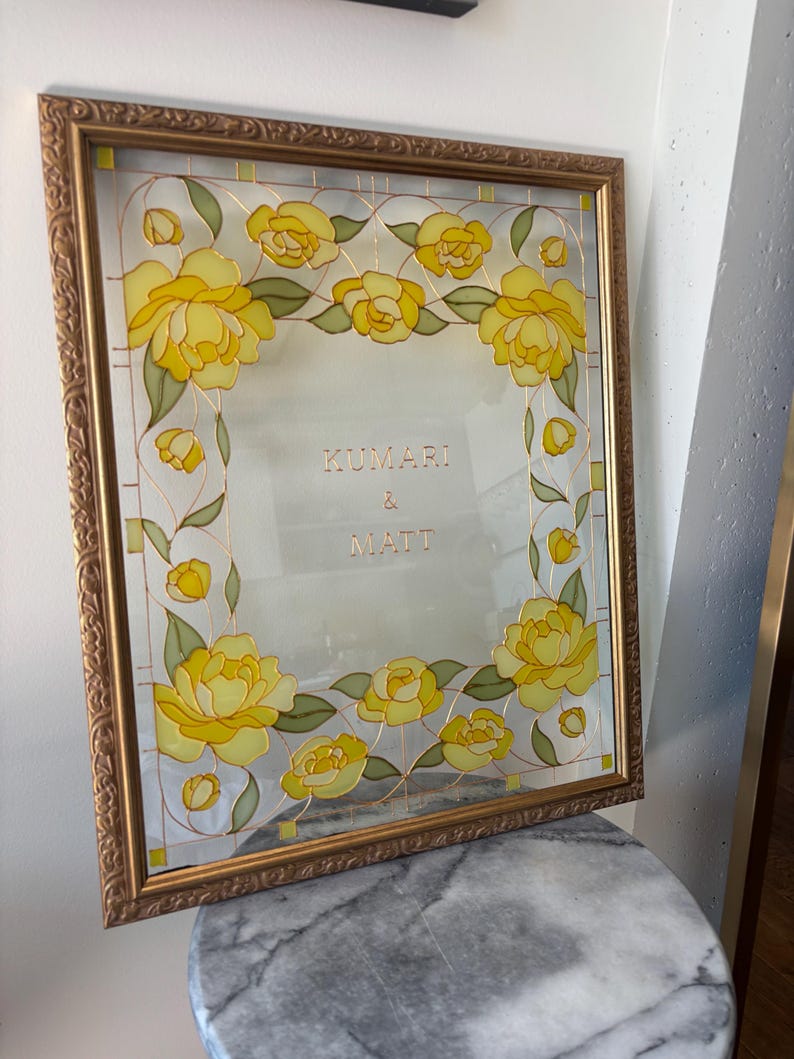 May include: A framed glass art piece with a gold-colored frame. The glass features a floral design with yellow roses and green leaves. The names "KUMARI & MATT" are displayed in gold lettering within the frame.