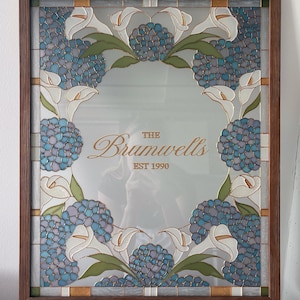 May include: A rectangular mirror in a dark wood frame featuring a stained glass border of blue hydrangea clusters, white calla lilies, and green leaves. The center of the mirror displays the text "THE Brumwells EST 1990" in gold lettering.