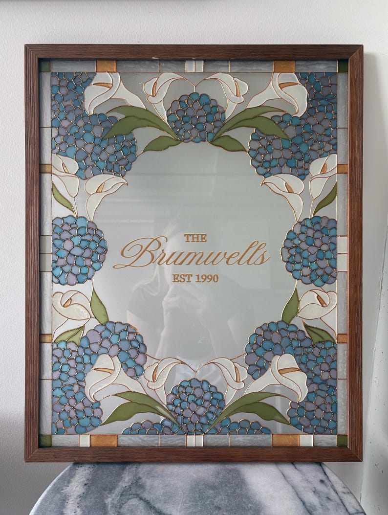 May include: A rectangular mirror in a dark wood frame featuring a stained glass border of blue hydrangea clusters, white calla lilies, and green leaves. The center of the mirror displays the text "THE Brumwells EST 1990" in gold lettering.