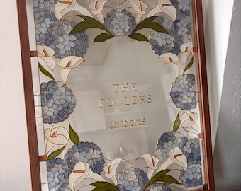 May include: A rectangular stained glass panel in a brown wooden frame featuring a floral border of blue hydrangea clusters and white calla lilies. The center is clear glass with the gold text THE FULLERS and the date 10.10.2026 etched in the middle.