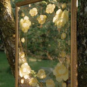 May include: A framed glass panel with a floral design and the names "Kumari & Matt" in gold lettering. The frame is gold-colored, and the flowers are yellow and white.