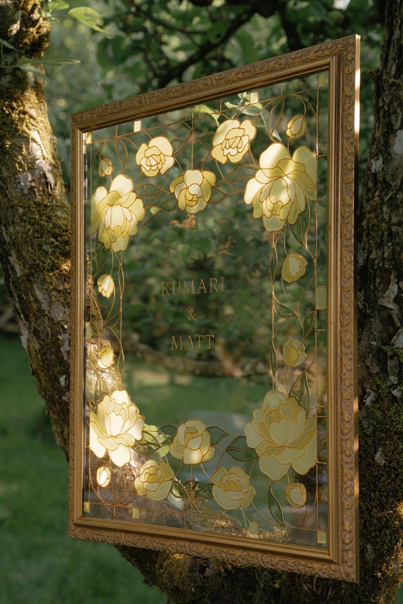 May include: A framed glass panel with a floral design and the names "Kumari & Matt" in gold lettering. The frame is gold-colored, and the flowers are yellow and white.