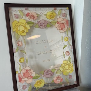 May include: A framed floral design with a dark brown wooden frame. The artwork features yellow, pink, and purple flowers with green leaves. The text "CHELSEA & CAMERON 06.12.26" is visible within the frame.