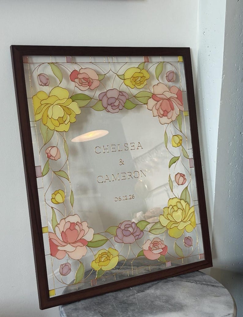 May include: A framed floral design with a dark brown wooden frame. The artwork features yellow, pink, and purple flowers with green leaves. The text "CHELSEA & CAMERON 06.12.26" is visible within the frame.