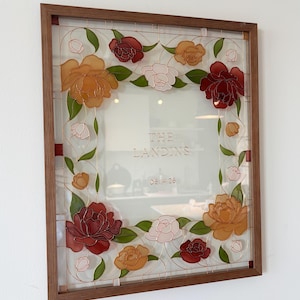 May include: A framed glass art piece featuring a floral design. The artwork showcases a square frame with a brown wooden border. The glass is adorned with colorful flowers in shades of red, orange, and pink, with green leaves. The text "THE LANDINS" and "08.14.26" are visible.