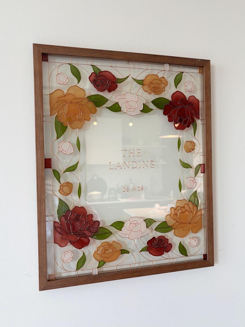 May include: A framed glass art piece featuring a floral design. The artwork showcases a square frame with a brown wooden border. The glass is adorned with colorful flowers in shades of red, orange, and pink, with green leaves. The text "THE LANDINS" and "08.14.26" are visible.