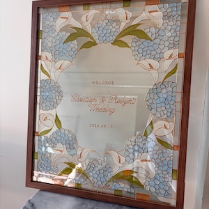 May include: A rectangular glass sign in a brown frame featuring a decorative border of light blue hydrangea flowers and white calla lilies. The center text reads: WELCOME, Christian & Robyn's Wedding, 2026.09.12, written in brown lettering.