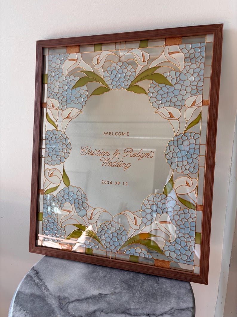 May include: A rectangular glass sign in a brown frame featuring a decorative border of light blue hydrangea flowers and white calla lilies. The center text reads: WELCOME, Christian & Robyn's Wedding, 2026.09.12, written in brown lettering.
