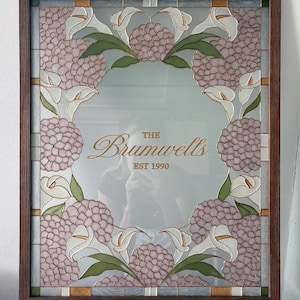May include: A rectangular mirror framed in dark wood featuring a decorative stained glass border of pink hydrangea clusters and white calla lilies with green leaves. The center of the mirror displays the gold text The Brumwells EST 1990.
