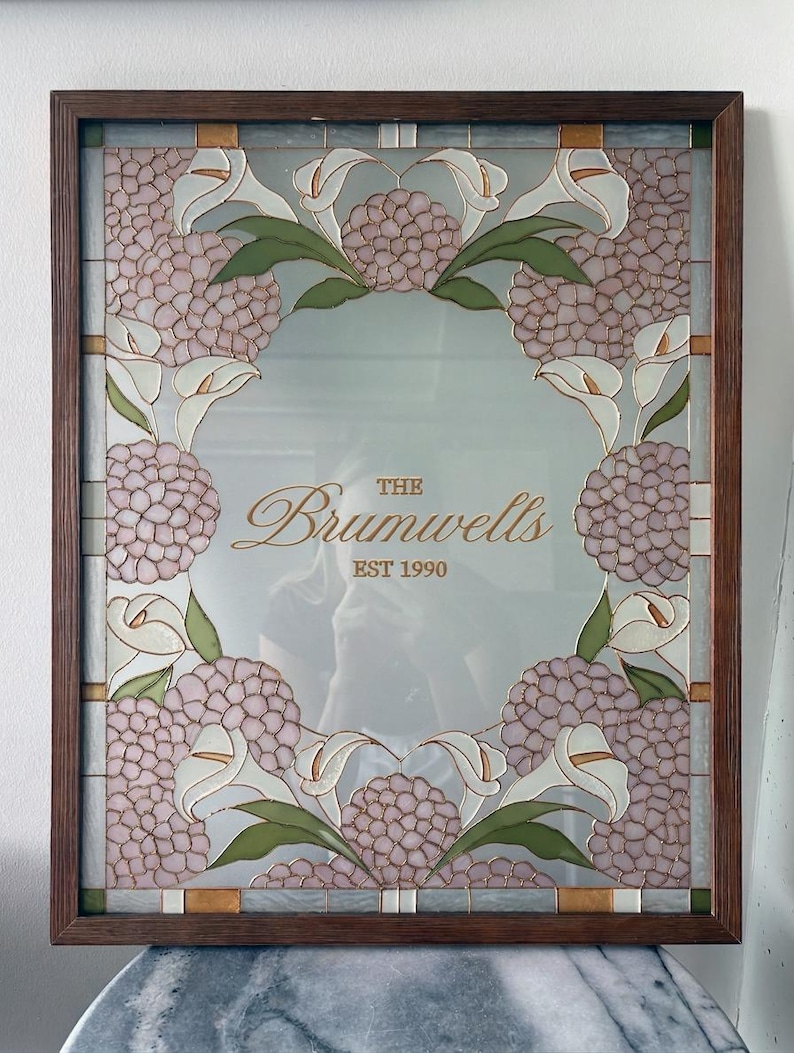 May include: A rectangular mirror framed in dark wood featuring a decorative stained glass border of pink hydrangea clusters and white calla lilies with green leaves. The center of the mirror displays the gold text The Brumwells EST 1990.