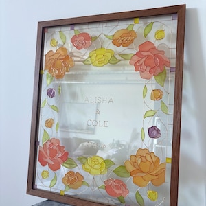 May include: A framed glass art piece with a floral design. The flowers are in shades of orange, yellow, and pink, with green leaves. The frame is a dark wood. The names "ALISHA & COLE" and the date "07.25.26" are visible.