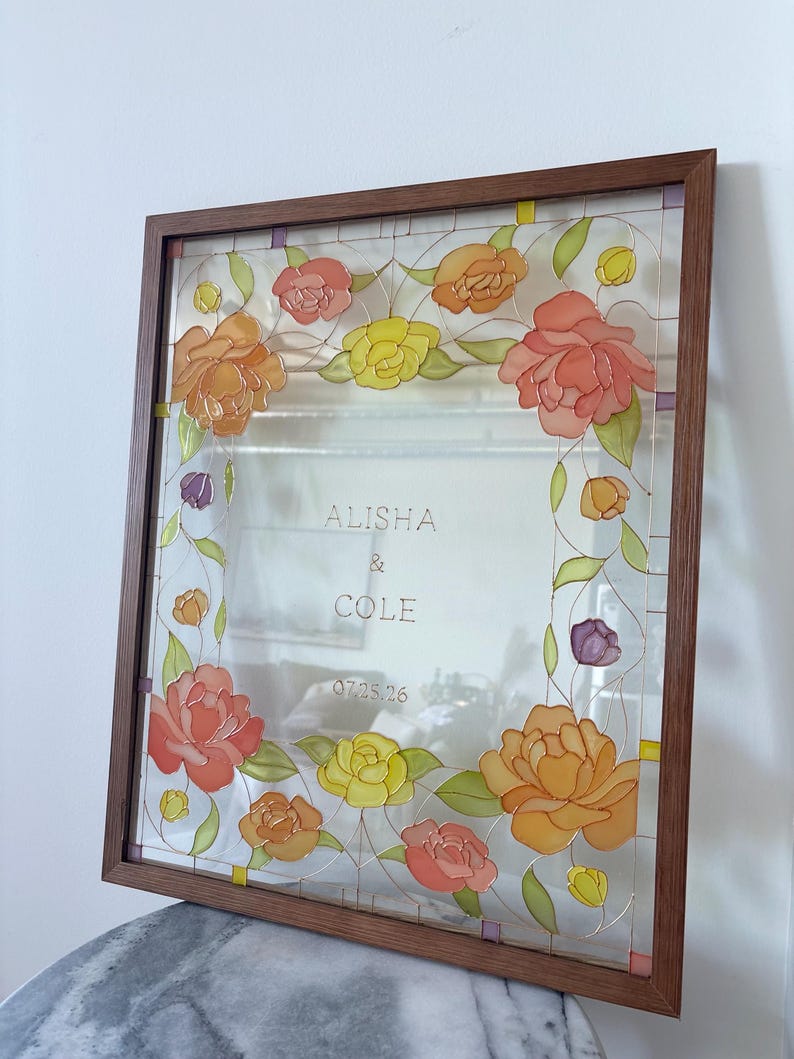 May include: A framed glass art piece with a floral design. The flowers are in shades of orange, yellow, and pink, with green leaves. The frame is a dark wood. The names "ALISHA & COLE" and the date "07.25.26" are visible.