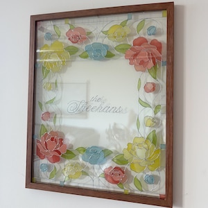 May include: A framed floral artwork featuring a brown wooden frame. The artwork showcases colorful flowers in shades of red, yellow, and blue, with the text "the Sheehans" in the center.