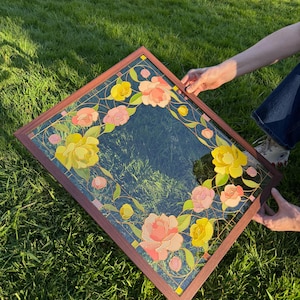 May include: A square decorative mirror with a dark blue background and a floral design. The flowers are in shades of yellow and pink, with green leaves and gold accents. The frame is a dark brown color.