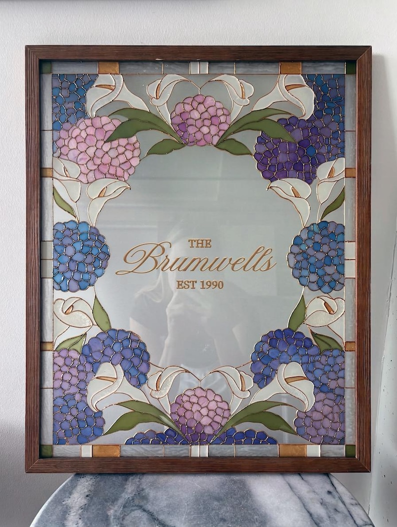 May include: A rectangular mirror framed in dark wood, featuring a decorative stained glass border of pink and blue hydrangea clusters and white calla lilies. The center of the mirror displays the gold text "THE Brumwells EST 1990".