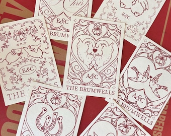 May include: A collection of rectangular cream-colored cards featuring maroon line art illustrations. Each card displays a central heart motif containing various symbols like animals, horseshoes, or names, with dates 09.26 and 2026 or 06.19.27.