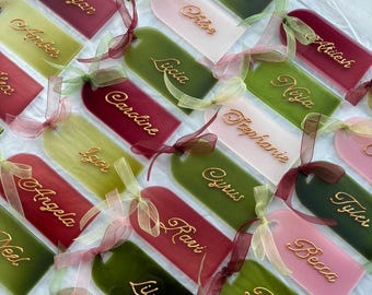 May include: A collection of rectangular acrylic tags with rounded tops, featuring various names in gold script lettering. Each tag is tied with a sheer ribbon in shades of green, pink, or burgundy, matching the tag color. The tags are arranged in a grid.