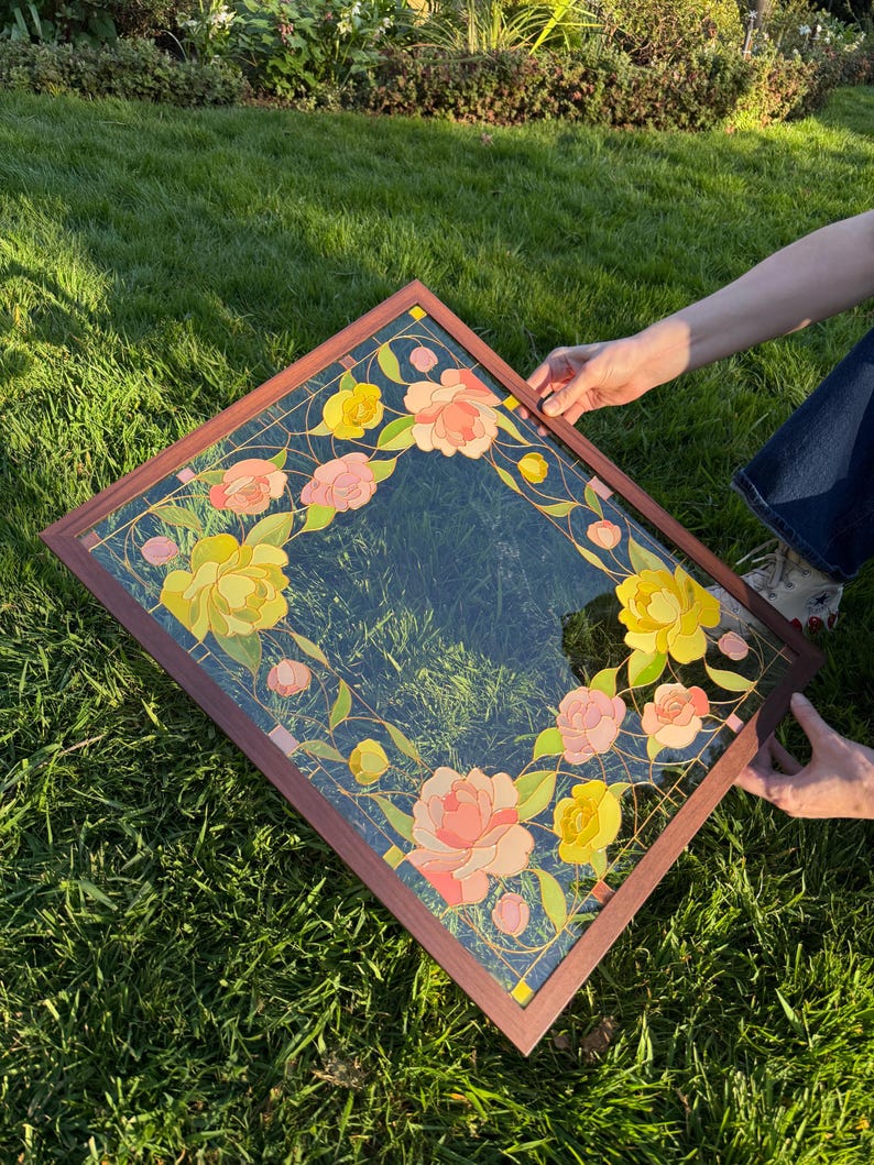 May include: A square mirror with a dark brown wooden frame. The mirror's surface is decorated with a floral design featuring yellow and pink flowers, green leaves, and gold accents. The mirror is held by two hands.