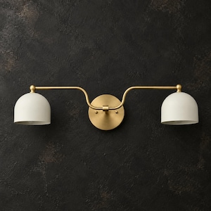 Puede incluir: Un aplique de pared de dos luces con una placa de montaje circular central de latón y dos brazos curvos de latón que se extienden hacia afuera, cada uno con una pantalla blanca mate en forma de cúpula orientada hacia abajo.