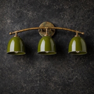 Puede incluir: Un aplique de pared de tres luces con una barra metálica curva de tono bronce texturizado y un soporte circular. Tres pantallas brillantes en forma de cúpula de color verde oliva cuelgan de la barra, cada una con un casquillo en su interior.