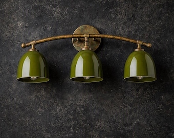 Può includere: Una lampada da parete a tre luci con una barra metallica curva color bronzo testurizzato e un supporto circolare. Tre paralumi lucidi a forma di cupola color verde oliva sono sospesi alla barra, ognuno con un portalampada all'interno.