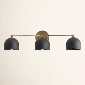 Puede incluir: Un aplique de pared de tres luces con pantallas en forma de cúpula de color negro mate unidas a una barra metálica horizontal en tono latón. La lámpara está montada sobre una placa circular de color latón detrás de la pantalla central.