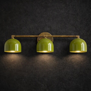 Puede incluir: Un aplique de pared con una barra horizontal de acabado bronce texturizado y tres pantallas brillantes en forma de cúpula de color verde oliva. Cada pantalla dirige la luz hacia abajo, creando un resplandor enfocado bajo las cubiertas.
