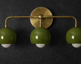 Puede incluir: Un aplique de pared con placa de montaje y brazo horizontal en acabado de latón cepillado. Tres pantallas brillantes en forma de cúpula de color verde oliva están unidas al brazo, cada una con una pequeña bombilla redonda de cristal blanco.