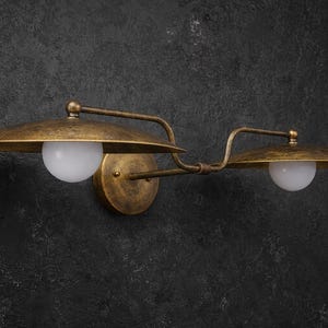 Puede incluir: Un aplique de pared con acabado en lat&oacute;n antiguo desgastado. Cuenta con dos brazos horizontales que se extienden desde una base circular central, cada uno sosteniendo una pantalla de metal ancha y plana con una bombilla de cristal esf&eacute;rica blanca.
