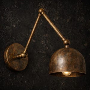 Puede incluir: Una lámpara de pared con un brazo articulado ajustable y una pantalla en forma de cúpula. El aplique tiene un acabado de metal bronce envejecido con una pátina oscura y texturizada. Se observa una bombilla dentro de la pantalla.