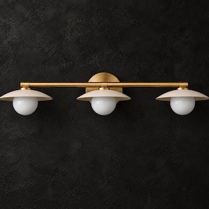 Puede incluir: Un aplique de pared de tres luces con una barra horizontal de latón y una placa circular de latón. Cada luz cuenta con una pantalla de metal blanco en forma de cúpula situada sobre una bombilla esférica de cristal blanco esmerilado.