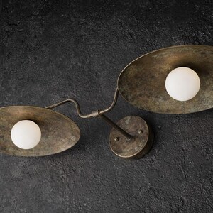 Puede incluir: Un aplique de pared con dos pantallas ovaladas de metal en tono bronce envejecido. Cada pantalla contiene una bombilla esf&eacute;rica de cristal blanco esmerilado. Las pantallas est&aacute;n unidas por un brazo met&aacute;lico curvo a una base circular.