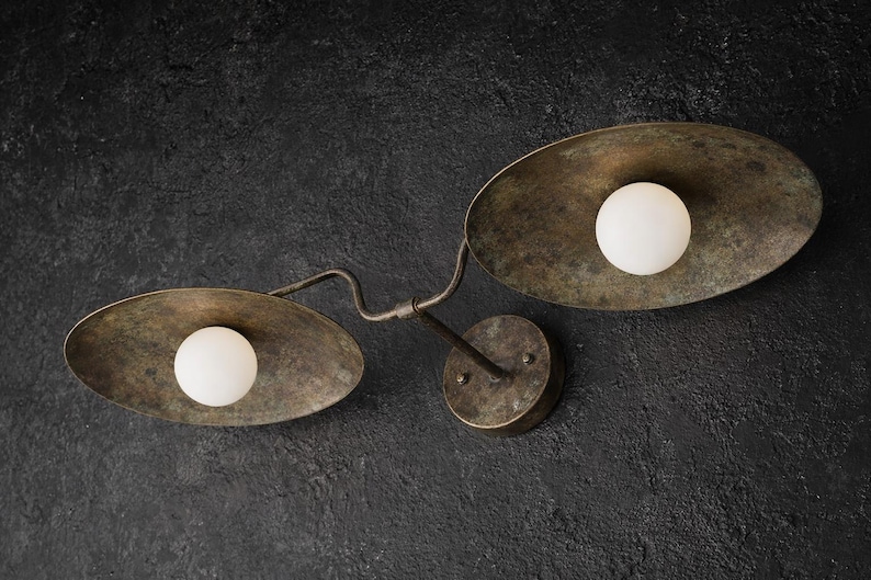 Puede incluir: Un aplique de pared con dos pantallas ovaladas de metal en tono bronce envejecido. Cada pantalla contiene una bombilla esf&eacute;rica de cristal blanco esmerilado. Las pantallas est&aacute;n unidas por un brazo met&aacute;lico curvo a una base circular.
