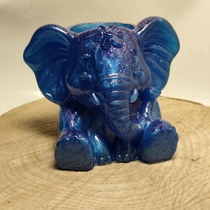 Puede incluir: Un recipiente azul con forma de elefante con detalles de purpurina. El recipiente está sobre una superficie de madera. El elefante tiene orejas grandes, una trompa y pequeños colmillos. Es probable que sea un objeto decorativo o un pequeño macetero.