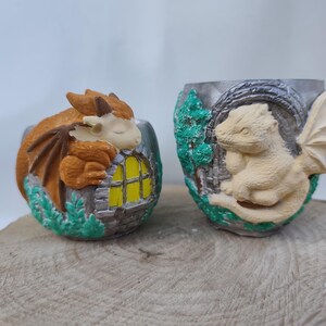 May include: Two decorative dragon-themed planters. One features a sleeping orange dragon with brown wings, nestled by a yellow-lit window. The other displays a cream-colored dragon with wings, set against a backdrop of green foliage and a stone wall.