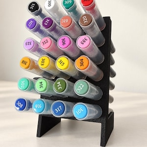 Op de afbeelding: Een zwarte displaystandaard met meerdere niveaus met vierentwintig transparante markers met gekleurde doppen. Elke dop is gelabeld met een specifiek nummer, waaronder 311, 300, 309, 305, 307, 313, 319, 320, 323, 312, 306, 355, 318, 303, 315, 304, 302, 314, 334, 322, 308, 916, 317 en 301.