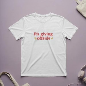 Puede incluir: Camiseta blanca con la frase "It's giving offside" en texto rojo y amarillo. La camiseta está centrada sobre un fondo morado, con una bolsa de mano y auriculares visibles.
