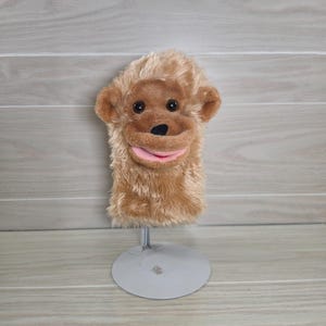 May include: A plush monkey hand puppet featuring light brown shaggy fur, dark brown plastic eyes, a black nose, and a pink mouth. The puppet is mounted on a silver-colored metal stand with a circular base, displaying the monkey's head and torso.