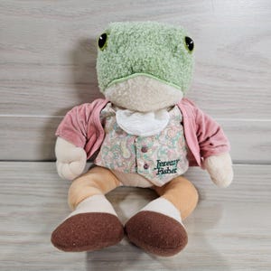 May include: A plush toy frog with a green textured head, large black eyes, and a cream-colored face. It wears a pink jacket over a paisley vest and a white ruffled collar. The text "Jeremy Fisher" is embroidered on the vest.