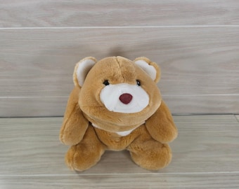 May include: A plush teddy bear with soft light brown fur, featuring a white muzzle, small black eyes, and a dark red nose. The stuffed animal has rounded ears with white inner accents and a white patch on its chest, presented in a seated position.