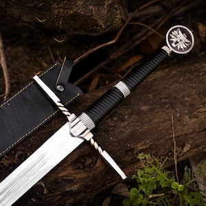May include: A silver-colored metal sword with a black ribbed handle and a circular pommel featuring a wolf head emblem. The sword has a twisted metal crossguard and is accompanied by a black leather scabbard with visible yellow stitching.