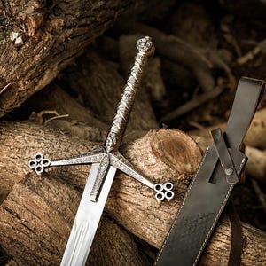 May include: A silver-toned metal sword featuring an intricate engraved hilt and a crossguard with four-lobed circular ends. The blade is straight and polished, accompanied by a dark brown leather scabbard with a matching strap and metal rivet.
