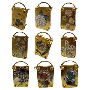 Peut inclure: Neuf poudriers en métal doré avec des motifs floraux et botaniques. Chaque poudrier a une forme rectangulaire, une petite poignée et des rivets décoratifs. Les motifs comprennent un trèfle à quatre feuilles, des pissenlits et diverses fleurs avec des accents argentés et colorés.