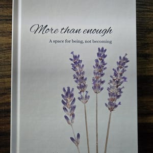 May include: A guided journal titled "More than enough" with lavender sprigs on the cover. The cover is light gray with the text in black script and a sans-serif font. The journal's subtitle reads "A space for being, not becoming."