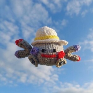 Op de afbeelding: Een gehaakt wezen met een grijze body, met een witte hoed met een gele band. Het heeft rode, paarse en zwarte accenten en een rode lachende mond. De achtergrond is een blauwe lucht met witte wolken.