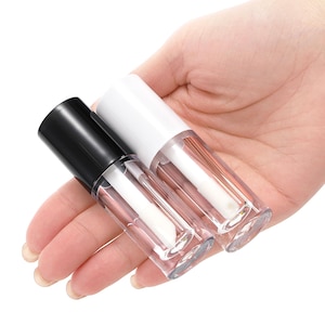 May include: Two empty lip gloss tubes with clear cylindrical bodies and applicator wands inside. One tube features a solid black cap, while the other tube features a solid white cap. The containers are transparent, showing the white applicator tips.