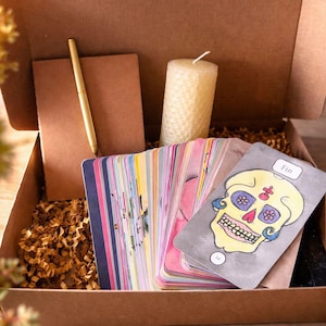Peut inclure: Coffret cadeau marron contenant divers articles. On y trouve un jeu de cartes colorées, une carte tête de mort jaune, un carnet marron, un stylo doré et une bougie texturée. Le coffret est garni de papier de soie brun.