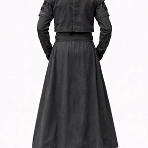 May include: A black, long-sleeved outfit with a cropped jacket and a long skirt. The jacket has shoulder accents and the sleeves have a subtle striped pattern. The skirt is full-length, and the outfit is belted at the waist. The pants are black.